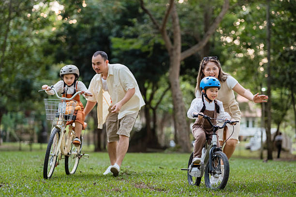 a family of four riding bikes in a park. a man is running to catch up with his children - safe & supervised environment child photos et images de collection